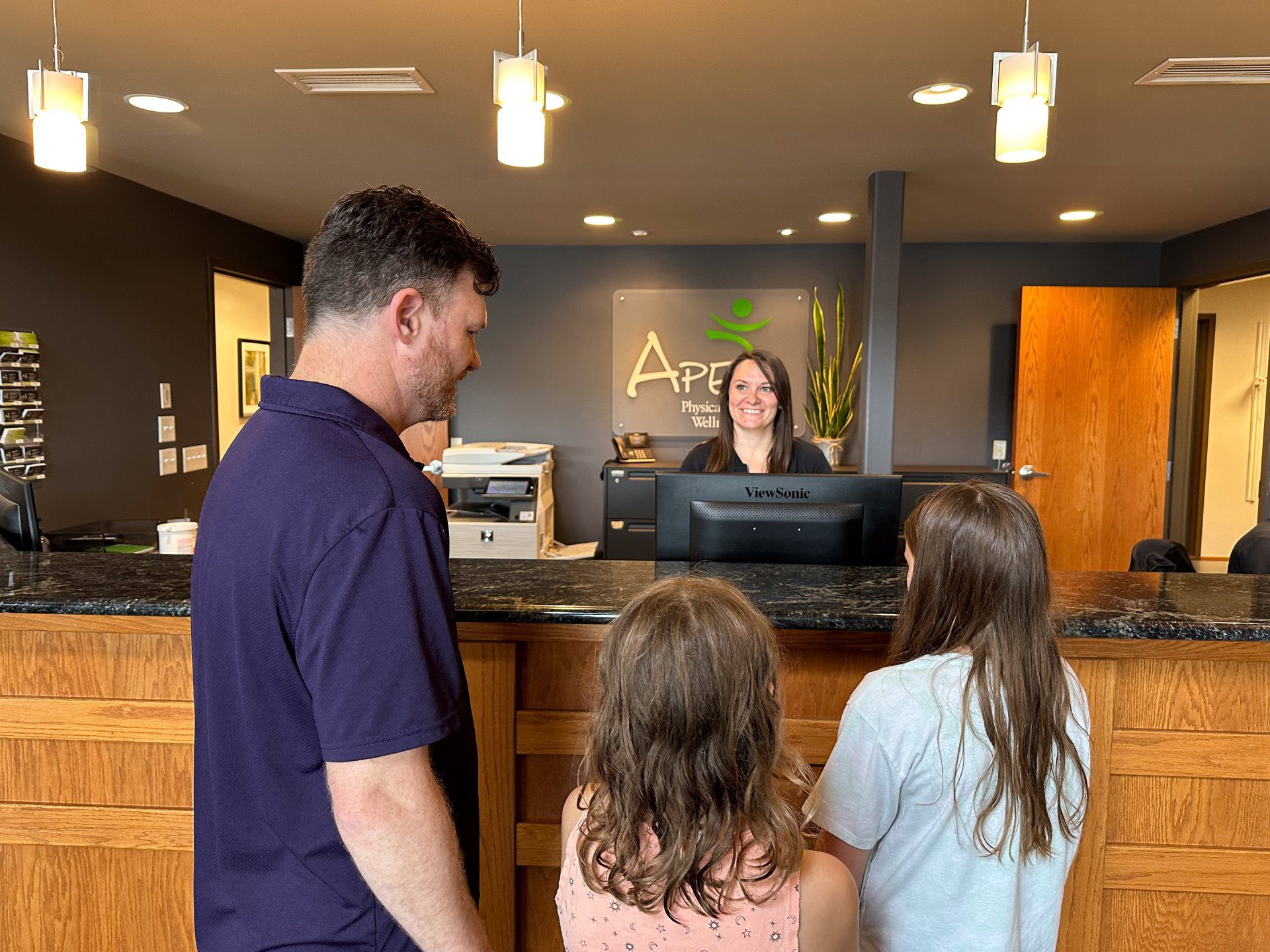 A man and two children approach the reception desk where a smiling receptionist, adhering to the Privacy Policy, awaits to assist them at a professional office.