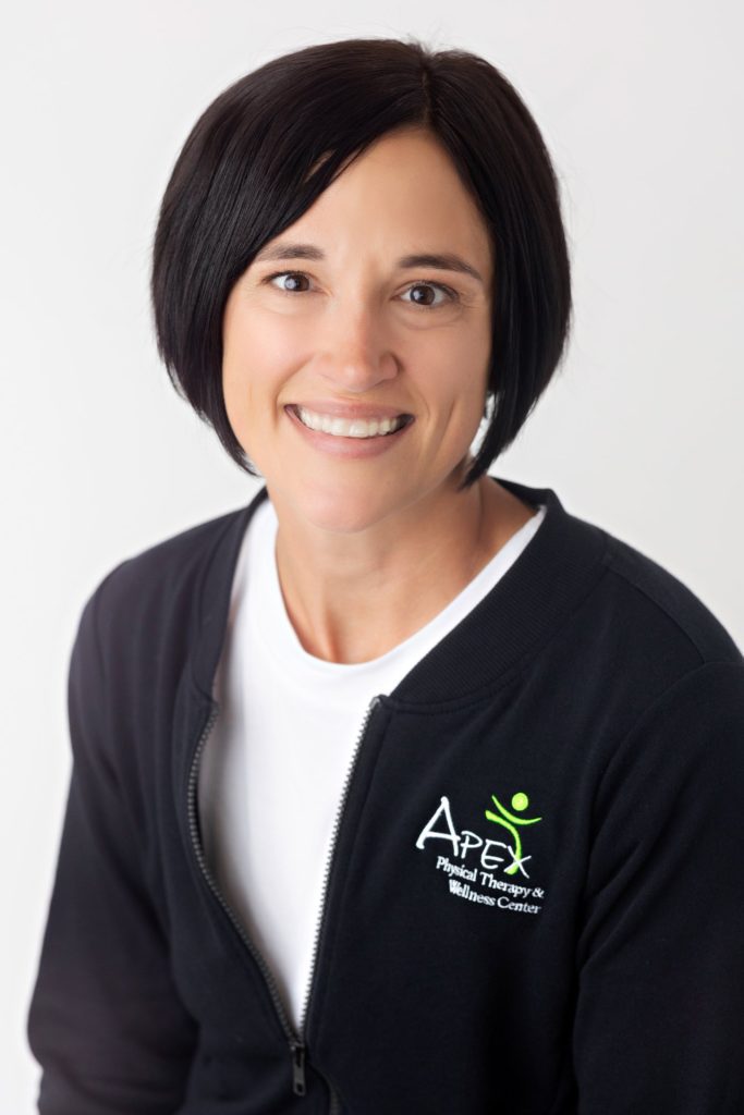 A professional portrait of Kristin Guderian, a smiling woman with short dark hair, wearing a black jacket with the logo "apex physical therapy & wellness center" embroidered on it, suggesting she
