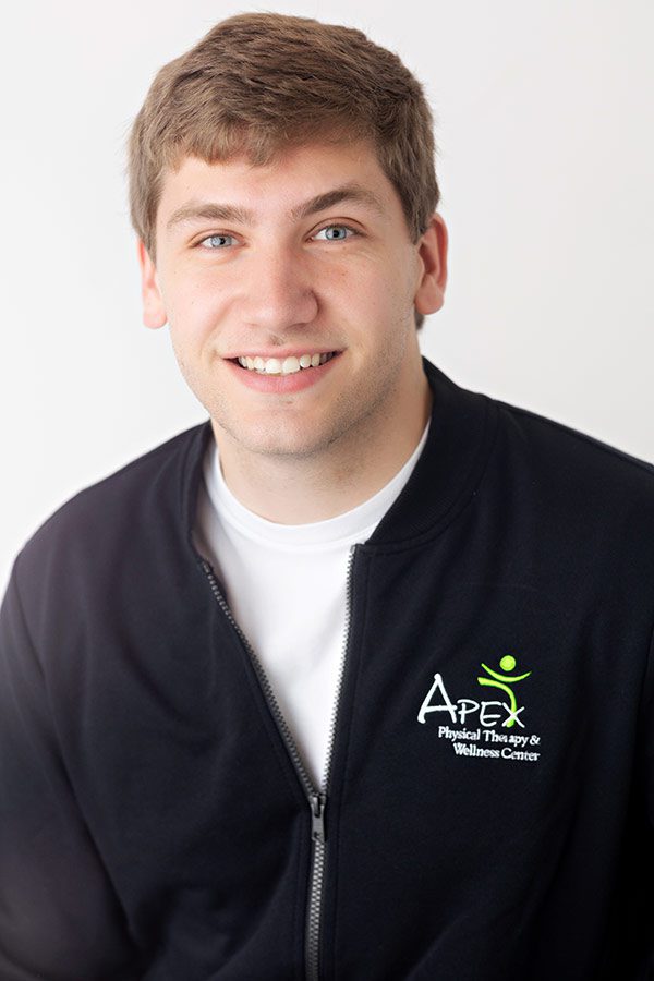 A young man smiling at the camera, wearing a black zip-up jacket with the Jonathan Tinklenberg physical therapy & wellness center logo on it.