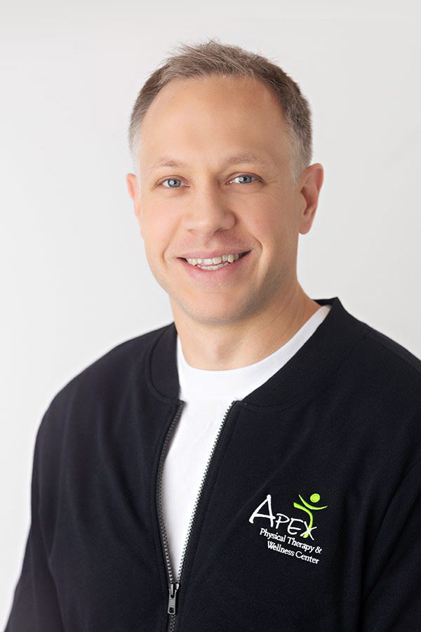 Professional headshot of a smiling man wearing a black zip-up jacket with an "apex physical therapy & wellness center" logo by Kayla Heger.