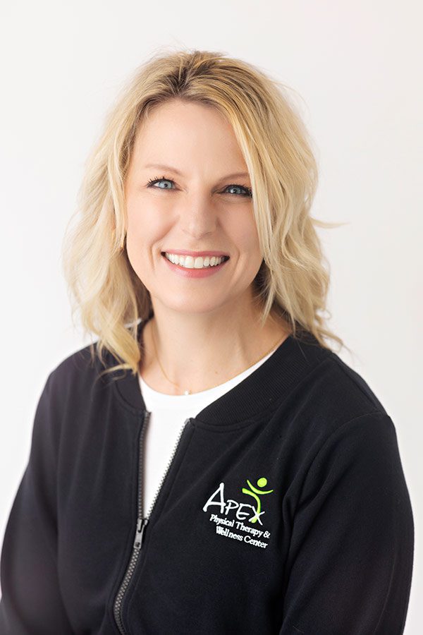A smiling professional woman wearing a black zip-up jacket with Jonathan Tinklenberg's Apex Physical Therapy & Wellness Center logo.