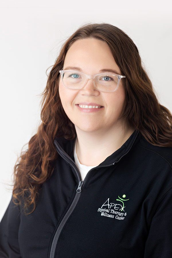 A smiling professional woman with glasses, wearing a black zip-up jacket with the Emma Neinaber Wellness and Therapy Center logo, against a light neutral background.