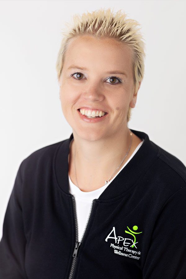 Professional portrait of a cheerful woman with spiky blonde hair, wearing a black zip-up jacket with the logo of Apex Physical Therapy & Wellness Center, captured by Jonathan Tinklenberg.