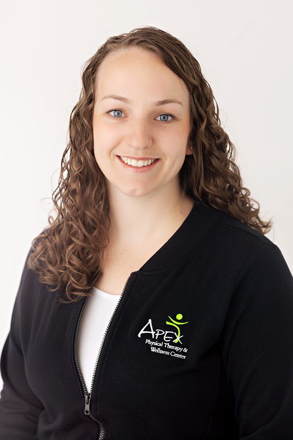 A smiling woman with curly hair, wearing a black zip-up jacket with the apex physical therapy & wellness center logo, posing for a professional portrait against a light background.