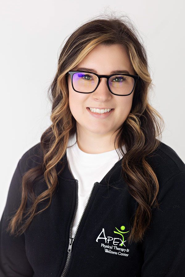 Casey Sailer, a friendly professional woman with wavy hair, wearing glasses and a black jacket with the apex physical therapy & wellness center logo, smiles warmly against a light background.