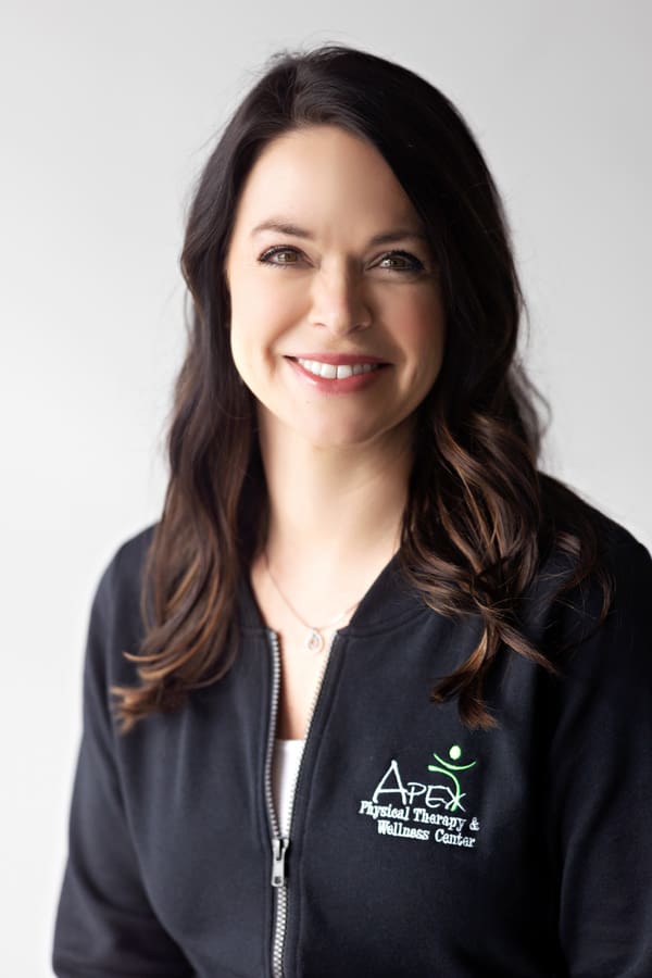 A confident professional woman with a friendly smile, wearing a black jacket with the logo of Apex Physical Therapy & Wellness Center, is Jessica Christenson.