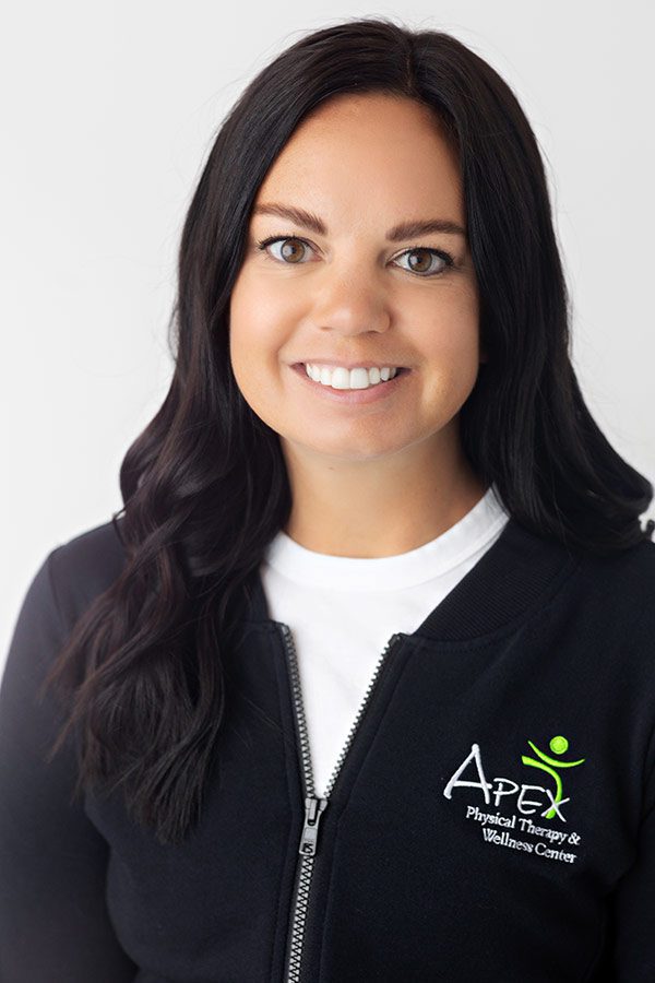 A professional headshot of Kayla Heger, smiling and wearing a black zip-up jacket over a white collared shirt, with the logo of a physical therapy and wellness center on her left chest.