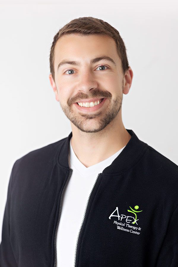 A cheerful man with a beard, dressed in a black zip-up jacket with an "apex physical therapy & wellness center" logo, posing against a plain white background is Josh Sorvig.