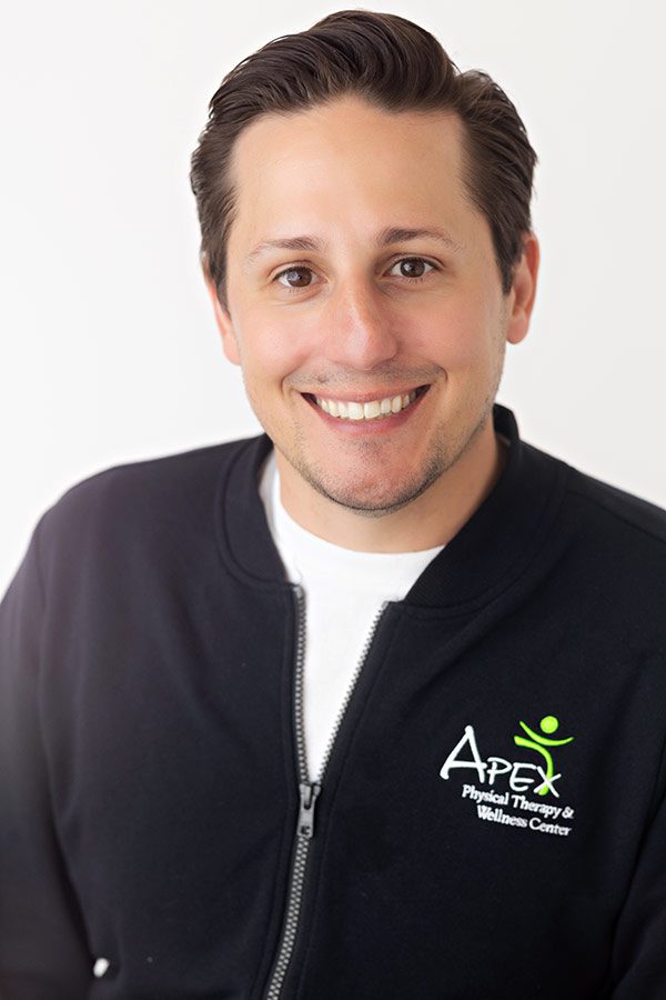 A friendly male healthcare professional named Kameron Ihry Hodem in a branded Apex Physical Therapy & Wellness Center jacket smiling for a portrait.