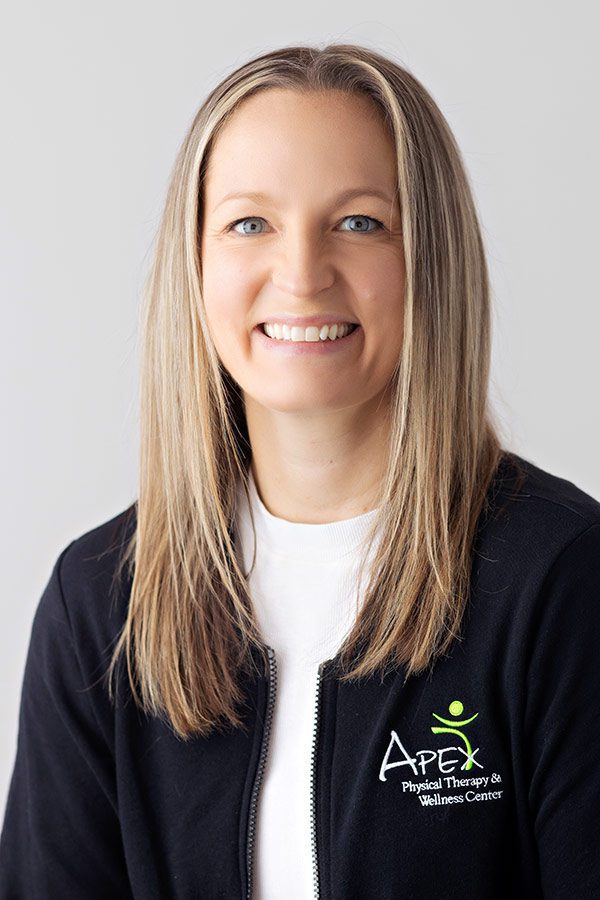 A smiling woman with long hair, wearing a black jacket with the "apex physical therapy & wellness center" logo, posing against a light gray background in a professional portrait featuring Casey Sailer.