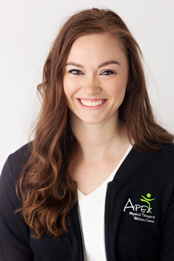 A professional woman named Kayla Heger, with long, wavy hair is wearing a black zip-up jacket with the "apex physical therapy & wellness center" logo, smiling brightly against a clean