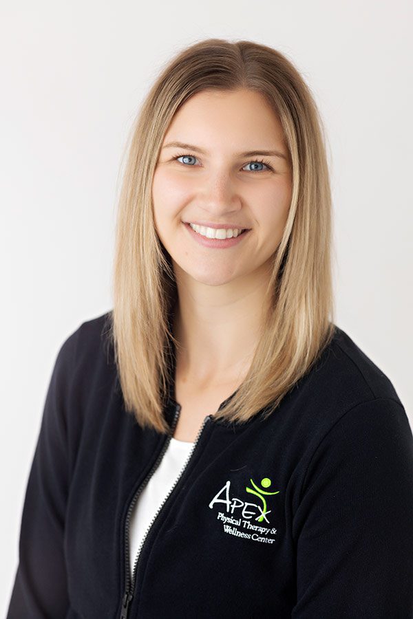 A smiling woman with shoulder-length blond hair, wearing a black jacket with the "Josh Sorvig Apex Physical Therapy & Wellness Center" logo, posing for a professional portrait against a light-colored background.
