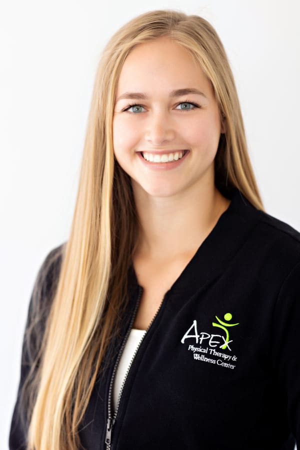 A professional portrait of Beth Prashek smiling and wearing a black zip-up jacket with an "apex physical therapy & wellness center" logo.