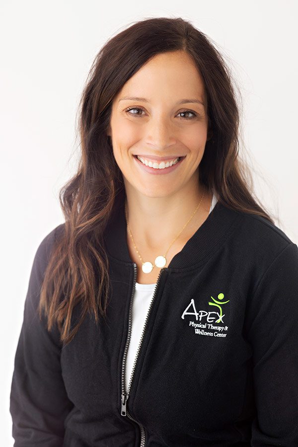 A professional woman with a friendly smile wearing a black jacket embroidered with the "Josh Sorvig Apex Physical Therapy & Wellness Center" logo.