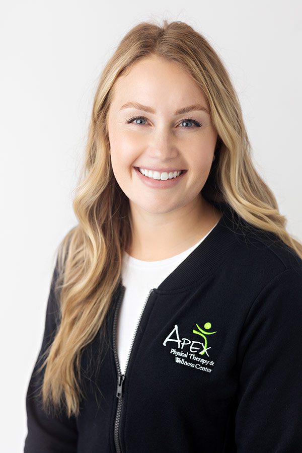A cheerful woman named Casey Sailer, wearing a black jacket with an apex physical therapy logo, is standing against a white background and offering a warm, friendly smile.
