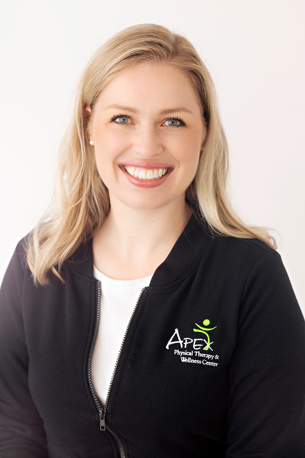 A smiling woman with blonde hair, wearing a black jacket with the Apex Physical Therapy & Wellness Center logo, poses against a light background.