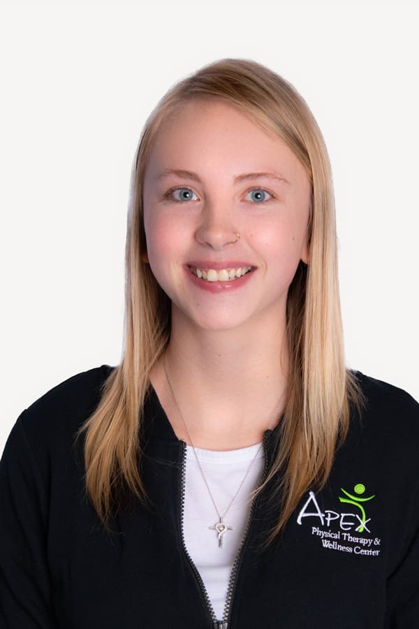 A smiling young woman with blonde hair, Beth Prashek, wearing a black zip-up jacket with the Apex Physical Therapy & Wellness Center logo, against a white background.