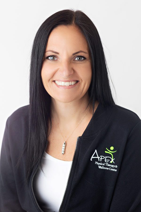 A professional woman named Casey Sailer, with long dark hair and a friendly smile, wearing a black jacket with the logo of Apex Physical Therapy & Wellness Center embroidered on it.