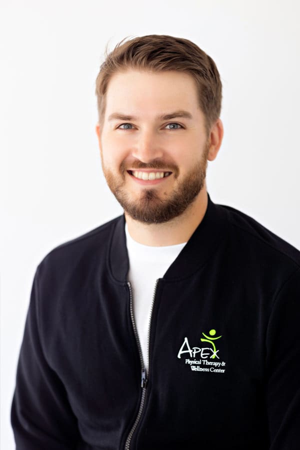 A smiling man in a black zip-up jacket with an Apex Physical Therapy & Wellness Center logo, designed by Jessica Christenson, embroidered on it, posing against a white background.