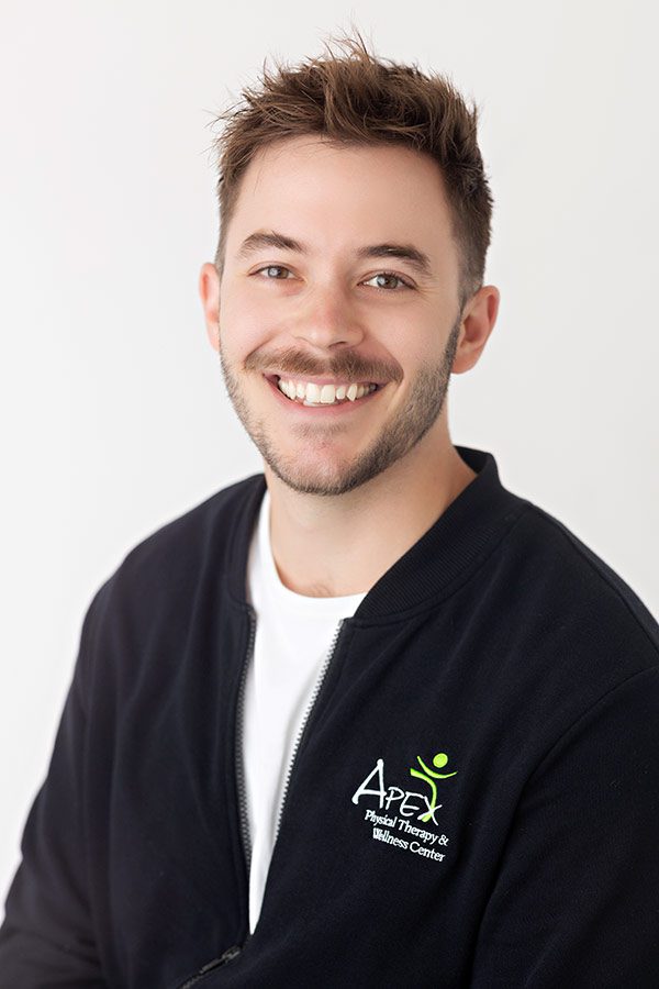 A cheerful young man with a stubble beard, wearing a black jacket with an embroidered logo, smiling confidently at the camera against a light neutral background.